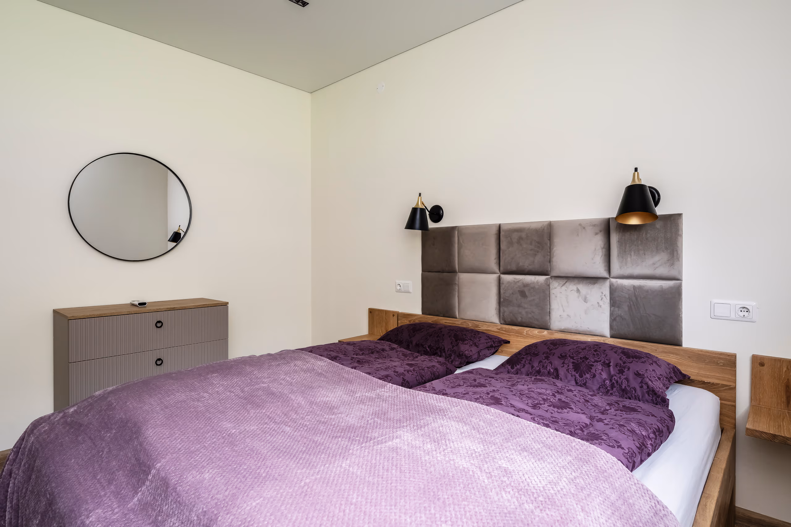 Bedroom with grey velvet headboard, black-brass wall sconces, lilac bedding and a round wall mirror
